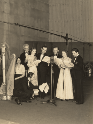 Group of male and female singers stand in front of microphones with woman playing the harp