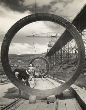 Men working on penstocks for Shasta dam; dam under construction at right with crane in place