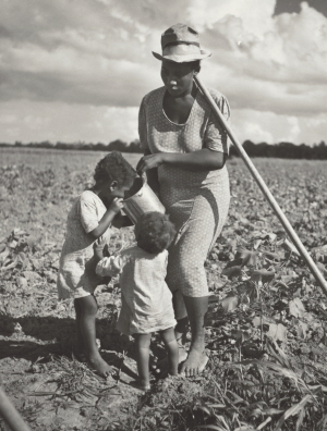 A woman stands barefoot in a field, a hoe resting against her shoulder. She holds a tin pail as one of her children takes a drink, the younger one waits for their turn.