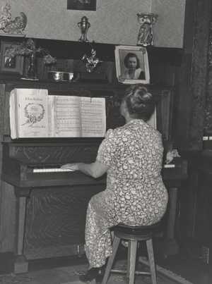 A woman plays an upright piano with her back turned away from the camera. The top of the piano is decorated with a trophy and two framed portraits.