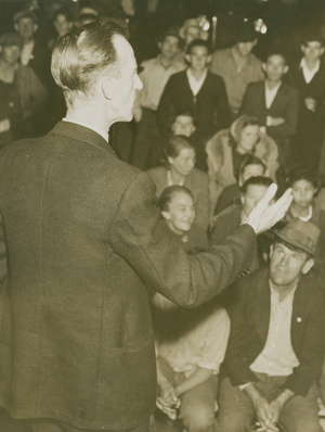 A man standing in front of a large group of workers gestures with his hands as he speaks.