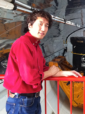 Portrait of Jonathan Jiang, standing in front of red railing. He is wearing a red shirt and blue jeans. Jet equipment can be seen in the background.