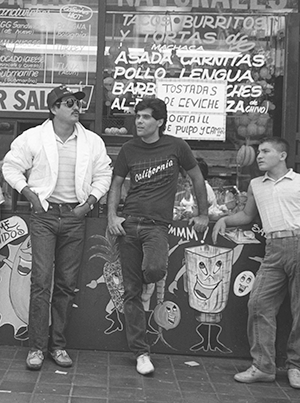 View of men leaning against food service counter on Broadway in Los Angeles. A food counter's menu signs can be seen behind them.