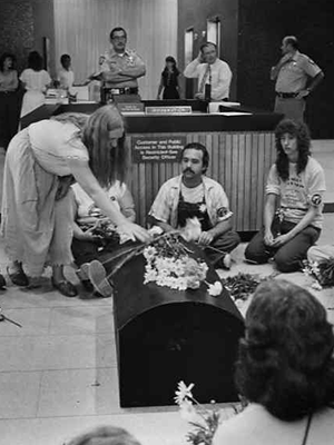 Image of people protesting the Rancho Seco Nuclear Power Plant. There is a small black coffin in the foreground, a woman placing a white flower on the coffin, and other protesters sitting on the floor. Security guards and other people can be seen behind a desk in the background.