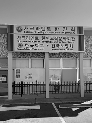 Black and white image of a building. A sign with text in English and Korean is hanging above windows.